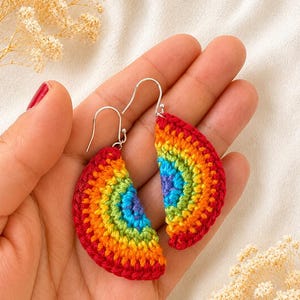 May include: A pair of semicircular crochet earrings featuring concentric bands of red, orange, yellow, green, blue, and purple yarn. Each earring is attached to a silver-toned metal hook, creating a vibrant rainbow-coloured accessory.