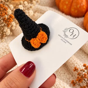 May include: A small black crocheted witch hat accessory featuring an orange crocheted bow at the base. The item is attached to a white display card printed with the text OH OZGUN HANDMADE STUDIO, featuring a circular logo with floral accents.