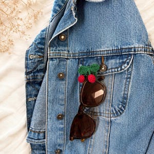 May include: A light blue denim jacket with brown sunglasses clipped to the pocket. A small, handmade pin with red pom-poms and a green bow is attached to the jacket. The jacket is buttoned up and has a classic design.