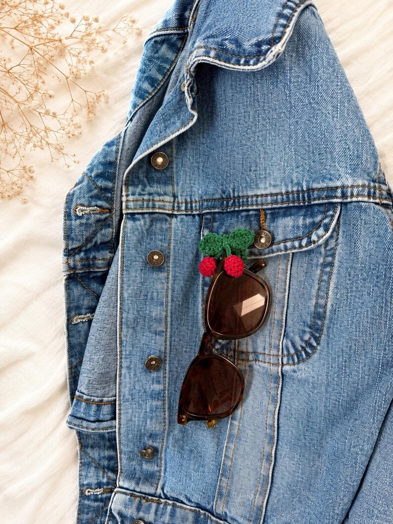 May include: A light blue denim jacket with brown sunglasses clipped to the pocket. A small, handmade pin with red pom-poms and a green bow is attached to the jacket. The jacket is buttoned up and has a classic design.