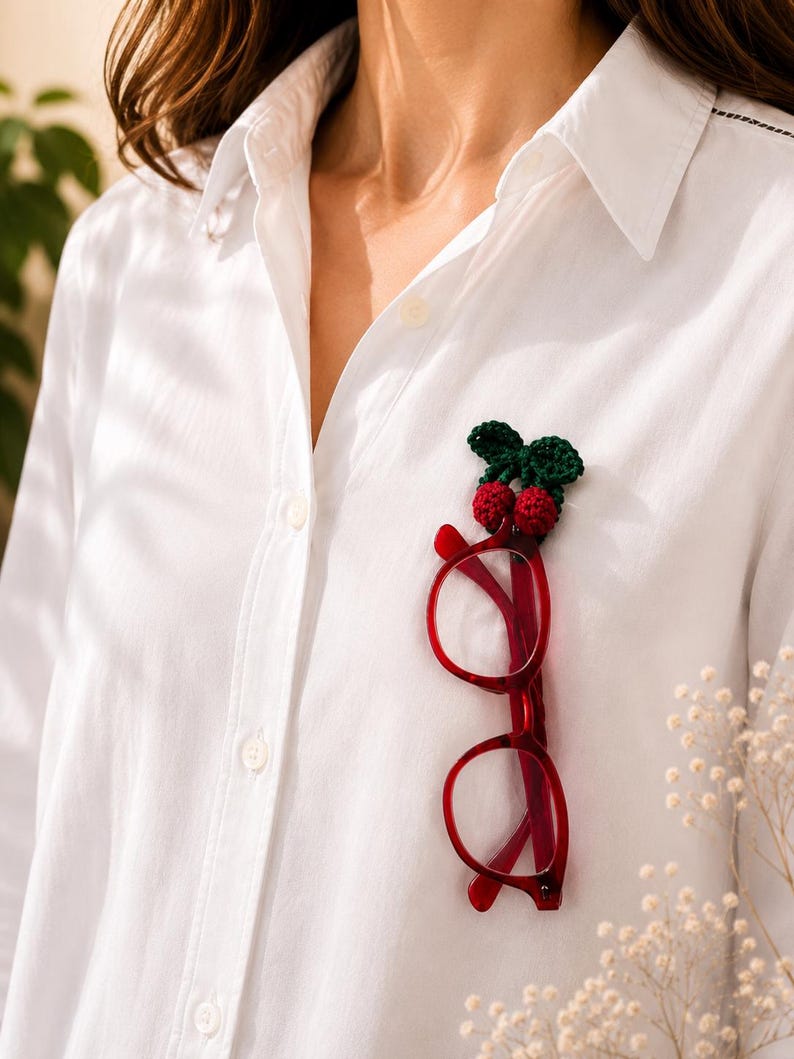 May include: A white shirt with a red-framed pair of spectacles clipped to it. A small, crocheted cherry brooch with green leaves is attached to the shirt. The spectacles have a tortoiseshell pattern.