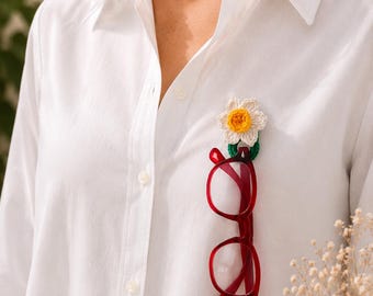 May include: A white shirt with a crochet flower brooch and red spectacles attached. The crochet flower has white petals and a yellow centre. The spectacles are a bright red colour.