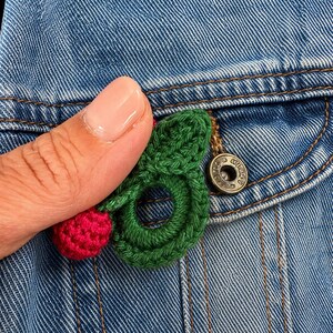 May include: A denim jacket pocket with a handmade crochet cherry pin. The pin has a red cherry and green leaves. The jacket has a metal button and visible stitching. The pocket is on a blue denim jacket.