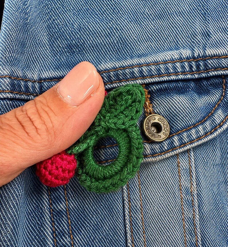 May include: A denim jacket pocket with a handmade crochet cherry pin. The pin has a red cherry and green leaves. The jacket has a metal button and visible stitching. The pocket is on a blue denim jacket.