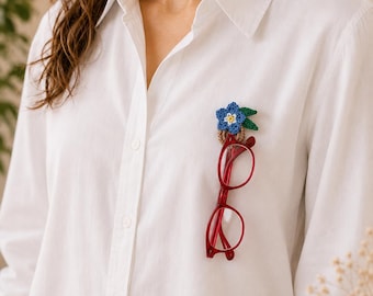 May include: A white shirt with a blue and green floral brooch and red spectacles attached. The brooch is a crocheted flower with blue petals, a yellow centre, and green leaves. The spectacles are a vibrant red colour.