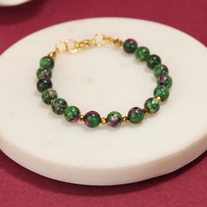 Pode incluir: Uma pulseira de contas com pedras redondas de zoisite verde e magenta intercaladas com pequenas contas espaçadoras de metal dourado. A pulseira é finalizada com um fecho lagosta de metal dourado e um anel de conexão circular.