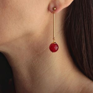 Peut inclure: Boucle d'oreille dorée avec une longue barre fine et une perle rouge facettée. La boucle d'oreille est présentée suspendue à une oreille, mettant en valeur le design et la couleur du bijou. La perle rouge est ronde et capte la lumière.