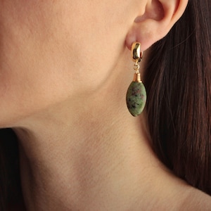 Puede incluir: Pendientes de aro dorados con una piedra verde ovalada colgante. La piedra tiene motas rojas y marrones. La piedra está sujeta al aro con alambre dorado. Los pendientes se muestran en la oreja de una persona.