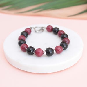 Peut inclure: Un bracelet composé de perles rondes noires et bordeaux alternées. Il est doté d'un fermoir à bascule argenté. Le bracelet est présenté sur une surface blanche et circulaire. Les perles mesurent environ 1 cm de diamètre.