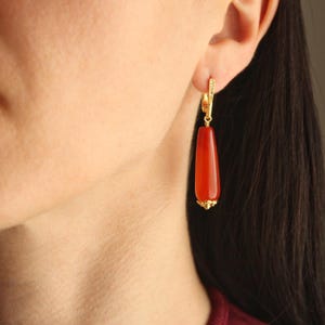 May include: A drop earring featuring a smooth, elongated, cylindrical red carnelian stone suspended from a gold-toned metal lever-back finding. The base of the stone is capped with a small, textured gold-toned metal decorative element.