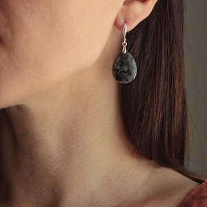 Puede incluir: Un solo pendiente en forma de lágrima con cierre plateado. El pendiente presenta una piedra gris oscuro con vetas grises y negras más claras. Se ve la oreja, el cuello y el pelo de la persona.