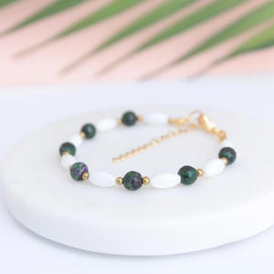 Op de afbeelding: Een armband met witte ovale kralen en donkergroene en rode ronde kralen. Het heeft een goudkleurige sluiting en ketting. De armband wordt gepresenteerd op een wit oppervlak.