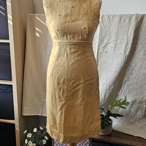 May include: A sleeveless mustard yellow dress featuring a round neckline and a fitted bodice. The upper portion of the dress is adorned with small, evenly spaced white buttons. The garment has a defined waistline and a straight skirt silhouette.