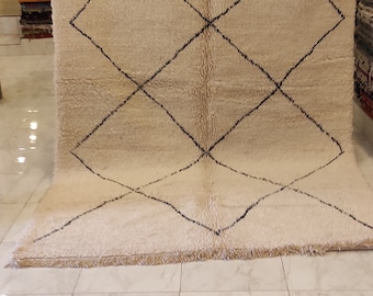 May include: Cream-colored, rectangular rug with a black diamond pattern. The rug has a thick, textured pile and a fringed edge. The rug is displayed in a shop setting, showcasing its design and texture. This rug is suitable for home decor.