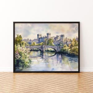 Op de afbeelding: Aquarel schilderij van een kasteel met meerdere torens en een stenen brug over een rivier. Het kunstwerk is ingelijst in een zwarte lijst. Weelderige groene bomen en bladeren omringen het kasteel en de brug, met reflecties in het water.