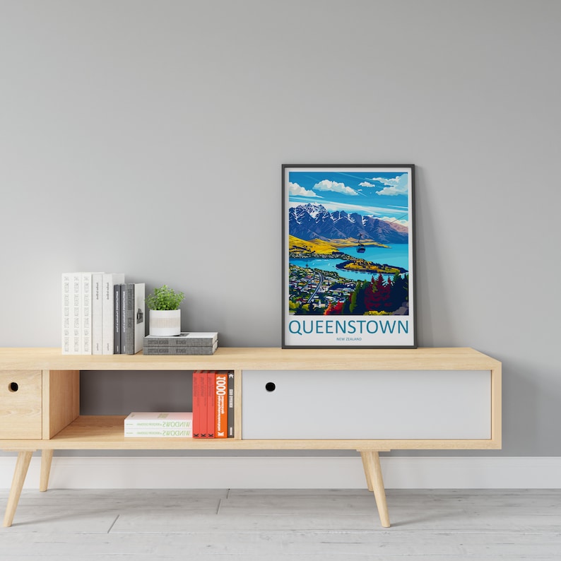 May include: A framed poster featuring a colourful illustration of Queenstown, New Zealand. The poster depicts a mountain range, a lake, and a gondola. The text "QUEENSTOWN, NEW ZEALAND" is printed at the bottom of the poster. The poster is displayed on a wooden shelf with books and a potted plant.