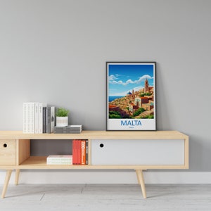 Puede incluir: Un p&oacute;ster enmarcado con una ilustraci&oacute;n colorida de una ciudad con un cielo azul y nubes blancas. El texto "MALTA" est&aacute; impreso debajo de la imagen. El p&oacute;ster est&aacute; en un estante de madera con libros y una planta en maceta.