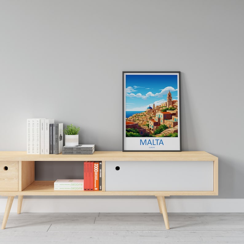 Puede incluir: Un p&oacute;ster enmarcado con una ilustraci&oacute;n colorida de una ciudad con un cielo azul y nubes blancas. El texto "MALTA" est&aacute; impreso debajo de la imagen. El p&oacute;ster est&aacute; en un estante de madera con libros y una planta en maceta.