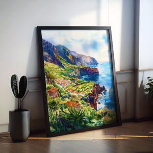 Pode incluir: Uma pintura em aquarela emoldurada de uma paisagem costeira com uma vila. A obra apresenta uma cena vibrante de colinas verdes em terra&ccedil;os, casas com telhados vermelhos e um oceano azul sob um c&eacute;u nublado. A moldura preta est&aacute; encostada em uma parede branca.