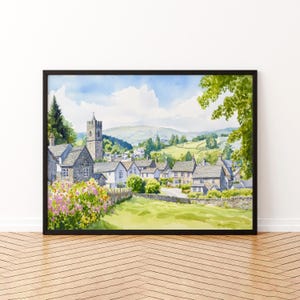 May include: A framed watercolor painting of a quaint village scene. The artwork depicts stone buildings, a church tower, and rolling green hills under a blue sky with fluffy clouds. Pink and yellow flowers bloom in the foreground.