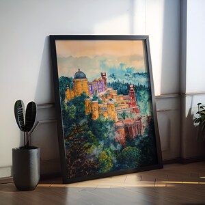 May include: Watercolor painting of a colorful castle on a hillside, framed in black. The artwork depicts a vibrant scene with yellow, purple, and red buildings surrounded by lush green trees and a blue sky. The framed art is leaning against a white wall.