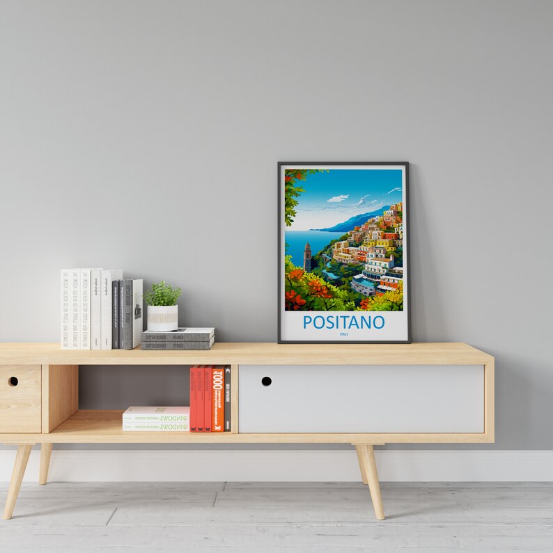 Puede incluir: Un p&oacute;ster enmarcado con una ilustraci&oacute;n colorida de Positano, Italia. El p&oacute;ster est&aacute; colgado en una pared sobre un estante de madera con libros y una planta en maceta.