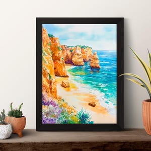 May include: Framed watercolor painting of a coastal landscape. The artwork depicts orange cliffs, a sandy beach, and turquoise water under a blue sky. The painting is in a black frame, displayed on a wooden surface with potted plants.