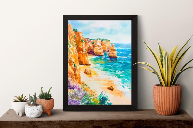 May include: Framed watercolor painting of a coastal landscape. The artwork depicts orange cliffs, a sandy beach, and turquoise water under a blue sky. The painting is in a black frame, displayed on a wooden surface with potted plants.