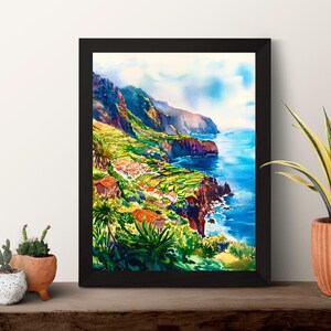 Pode incluir: Uma pintura em aquarela emoldurada retrata uma paisagem costeira com fal&eacute;sias, edif&iacute;cios e o oceano. A obra de arte apresenta cores vibrantes, incluindo azuis, verdes e laranjas. A pintura &eacute; exibida em uma superf&iacute;cie de madeira com plantas em vasos.