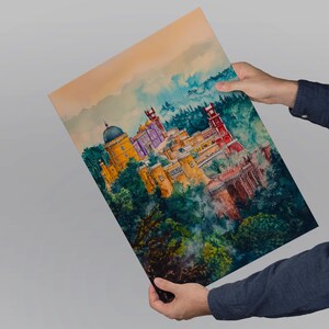 May include: A watercolor print of a colorful palace nestled in lush green trees. The artwork features a yellow dome, red towers, and purple accents. The print is held by a person wearing a dark blue long-sleeved shirt.