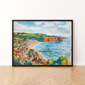 May include: Watercolor painting of a coastal town with red-roofed buildings, a sandy beach, and a pier. The artwork features a vibrant blue ocean, green hills, and a bright sky with fluffy clouds. The painting is framed in a black border.