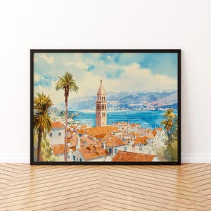 Könnte beinhalten: Aquarellmalerei einer Küstenstadt mit roten Ziegeldächern, weißen Gebäuden und einem hohen Glockenturm. Palmen umrahmen den Blick auf das blaue Meer und die fernen Berge. Das Kunstwerk ist in einem schwarzen Rahmen ausgestellt.