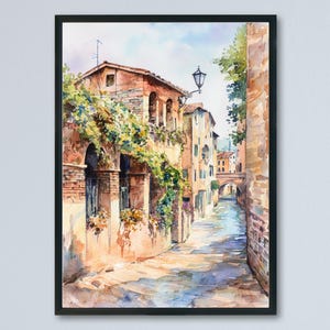 May include: Watercolor painting of a European street scene in a black frame. The artwork depicts a narrow street with buildings on either side, a canal, and a bridge in the distance. The buildings are in shades of orange, brown, and yellow.