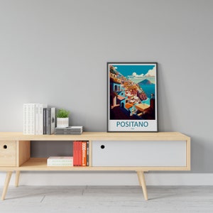 Może przedstawiać: Ramka z plakatem przedstawiającym kolorową ilustrację Positano we Włoszech. Plakat znajduje się na drewnianej p&oacute;łce z książkami i doniczką z rośliną.