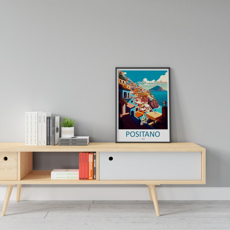 Może przedstawiać: Ramka z plakatem przedstawiającym kolorową ilustrację Positano we Włoszech. Plakat znajduje się na drewnianej p&oacute;łce z książkami i doniczką z rośliną.