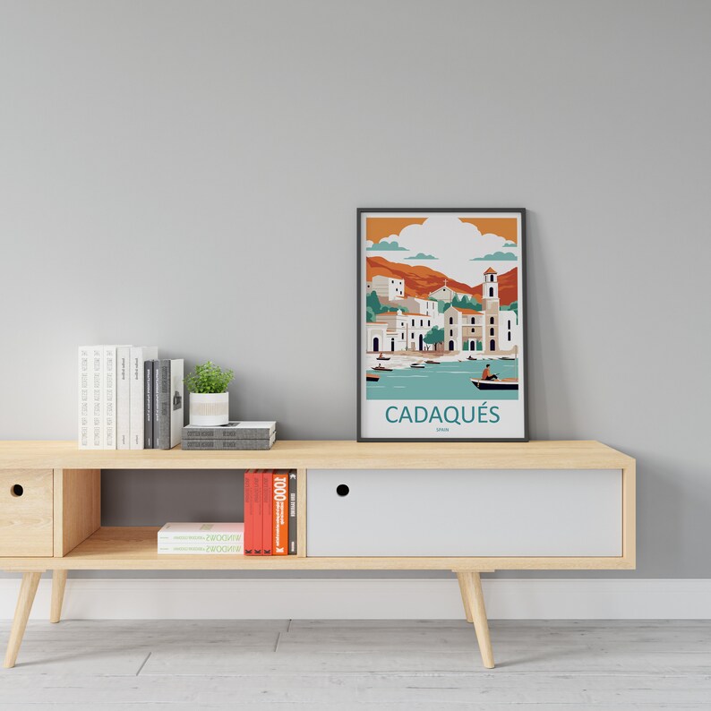 May include: A framed illustration of the town of Cadaqu&eacute;s, Spain, with a blue and green color scheme. The illustration depicts a coastal town with buildings, a church, and boats in the water. The frame is black and the text "Cadaqu&eacute;s, Spain" is printed below the illustration.
