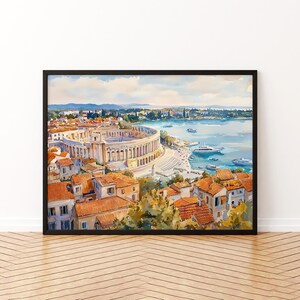 Puede incluir: Pintura a la acuarela de un pueblo costero con un gran anfiteatro antiguo. La obra presenta edificios con tejados de tejas naranjas, árboles verdes y un mar azul con barcos. La pintura está enmarcada en un marco negro.