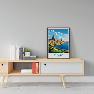 Puede incluir: Un p&oacute;ster enmarcado con una ilustraci&oacute;n colorida de una ciudad con una masa de agua azul y el texto "MALTA, EUROPA" en la parte inferior. El p&oacute;ster est&aacute; en un estante de madera clara con libros y una planta en maceta.
