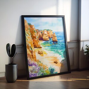 May include: Framed watercolor painting of a coastal landscape. The artwork depicts a beach with golden sand, turquoise water, and towering orange cliffs. The painting is in a black frame and leans against a white wall.