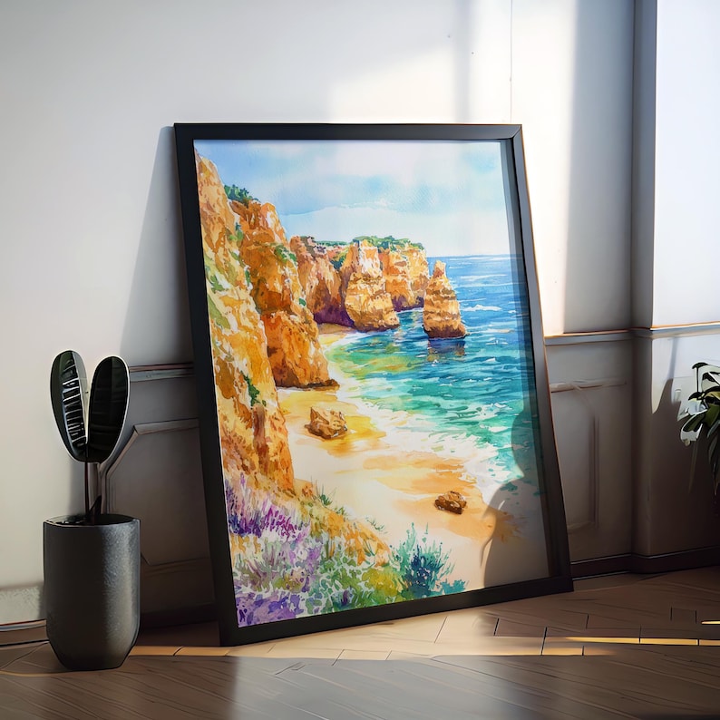 May include: Framed watercolor painting of a coastal landscape. The artwork depicts a beach with golden sand, turquoise water, and towering orange cliffs. The painting is in a black frame and leans against a white wall.
