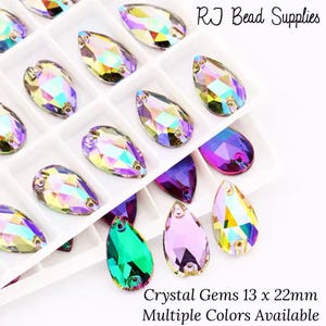 May include: Assortment of iridescent teardrop-shaped crystal gems in various colours, including purple, green, and pink. The gems are arranged in a white plastic tray with the text "RJ Bead Supplies" and "Crystal Gems 13 x 22mm Multiple Colours Available".