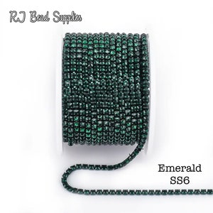 Peut inclure: Un rouleau de chaîne de strass vert émeraude. La chaîne est composée de petits strass carrés rapprochés. Le texte "Emerald SS6" est visible à droite de l'image. La marque "RJ Bead Supplies" est en haut.