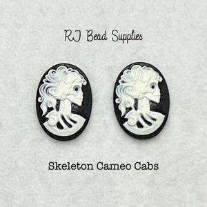 May include: Two black and white cameo cabochons featuring a skeleton design. The cabochons are oval shaped and have a white skeleton design on a black background. The text "Skeleton Cameo Cabs" is printed below the cabochons.