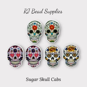 Peut inclure: Six cabochons en forme de crâne de sucre colorés avec différents motifs, y compris des fleurs, des cœurs et des motifs géométriques. Les cabochons sont blancs avec des contours noirs et présentent des couleurs vives comme le rose, le jaune, le bleu et le vert. Le texte "RJ Bead Supplies" et "Sugar Skull Cabochons" est visible sur l'image.