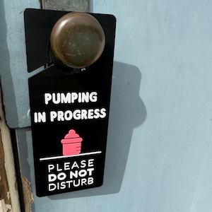 Puede incluir: Colgador de puerta negro con texto blanco que dice "Pumping in Progress" y "Please Do Not Disturb". Un icono de biberón rosa se encuentra en el centro del letrero.
