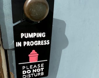 Può includere: Appendino per porta nero con testo bianco che recita "Pumping in Progress" e "Please Do Not Disturb". Un'icona di biberon rosa si trova al centro del cartello.