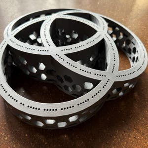 May include: A 3D-printed sculpture featuring three interlocking rings. The rings are black with a honeycomb pattern and are edged with a lighter gray color. Small dots are evenly spaced along the edges.