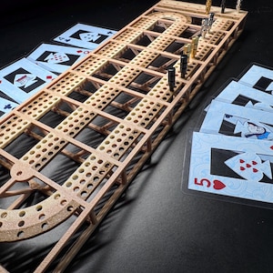 Può includere: Un tabellone di cribbage in legno con una pista di fori e un'estremità curva. Il tabellone ha pioli di vari colori. Le carte da gioco con un design blu e bianco sono sparse intorno al tabellone.