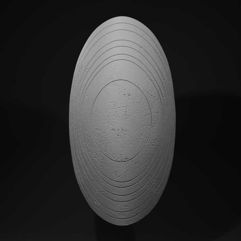 May include: A gray, oval-shaped shield with concentric circular grooves and a textured surface. The shield has a central circular design and is set against a dark background.