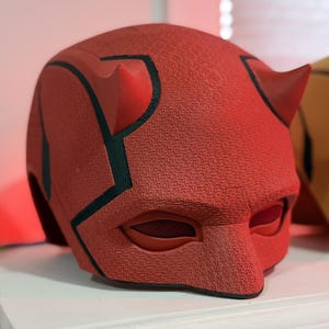 Może przedstawiać: Czerwono-czarna maska Daredevila z rogami. Maska ma fakturowaną powierzchnię i czarną maskę na oczy.